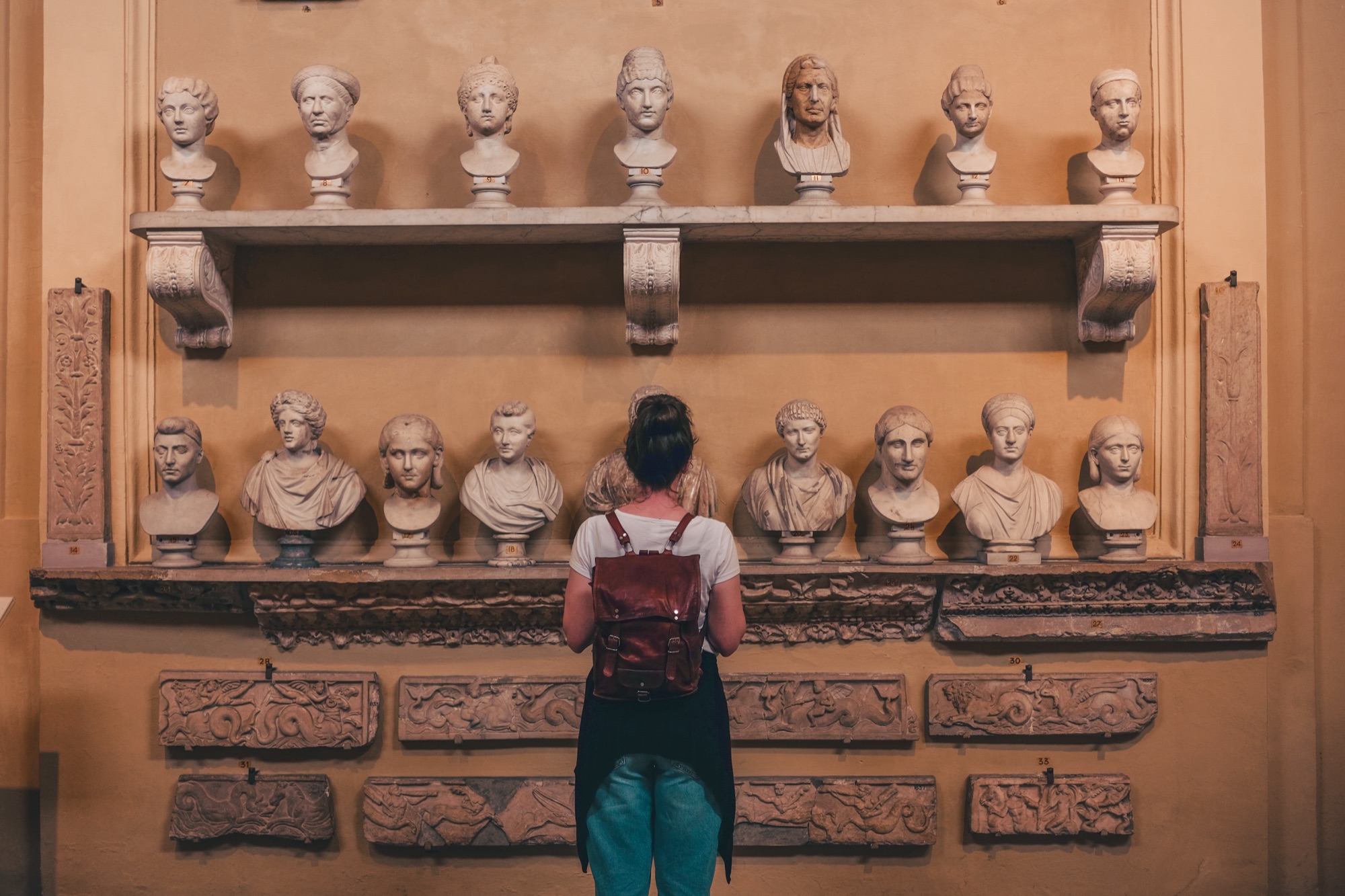 A visitor with a backpack standing before a wall of classical marble busts and carved reliefs in a museum